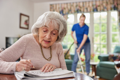 Elderly woman at home doing newspaper crossword while a younger female home services provider vacuums the floor in the background